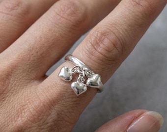May include: A silver ring featuring three dangling heart charms. The ring is worn on a finger, showcasing the delicate design and polished finish of the heart pendants. The ring's band is simple, allowing the hearts to be the focal point.