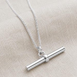 May include: A silver necklace featuring a T-bar pendant. The chain is a fine curb link design. The T-bar pendant is cylindrical with rounded ends and a small ring connecting it to the chain. The backdrop is a neutral beige fabric.