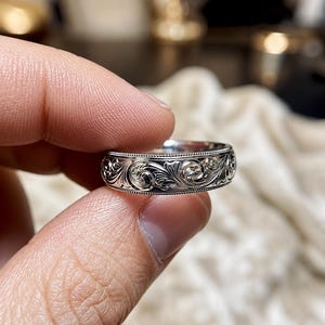 May include: A silver-toned metal ring featuring an intricate, continuous engraved scrollwork pattern across the outer band, bordered by fine beaded detailing along the top and bottom edges of the ring's circumference.
