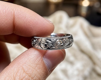 May include: A silver-toned metal ring featuring an intricate, continuous engraved scrollwork pattern across the outer band, bordered by fine beaded detailing along the top and bottom edges of the ring's circumference.
