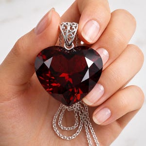 May include: A large, faceted, heart-shaped dark red gemstone pendant suspended from a silver-toned chain. The pendant features an intricate, openwork silver-toned metal bail with swirling filigree patterns at the top of the heart.