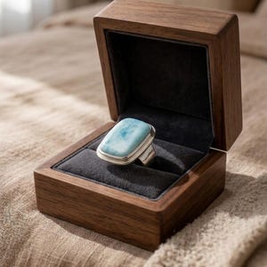 May include: A silver ring with a rectangular, light blue gemstone, presented in an open, dark wooden jewellery box with a black velvet lining. The box is on a textured surface. The ring is a statement piece.