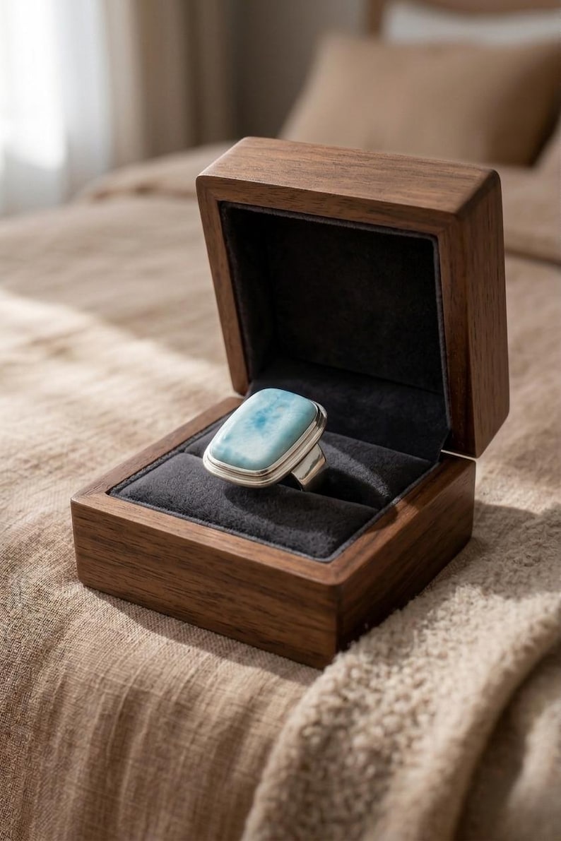 May include: A silver ring with a rectangular, light blue gemstone, presented in an open, dark wooden jewellery box with a black velvet lining. The box is on a textured surface. The ring is a statement piece.