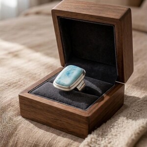 May include: A silver ring with a rectangular, light blue gemstone, presented in an open, dark wooden jewellery box with a black velvet lining. The box is made of dark wood.