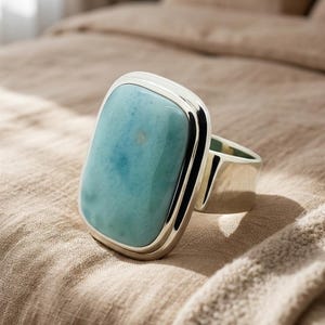 May include: A chunky ring featuring a rectangular, pale blue gemstone in a silver setting. The stone has a mottled pattern of blues and whites. The ring is presented on a neutral fabric, highlighting its bold design and size. The ring's design is modern.