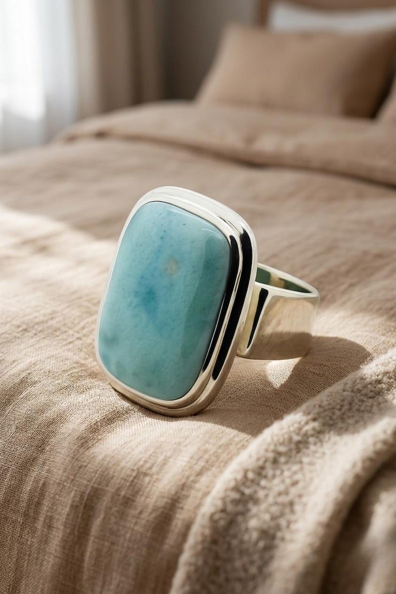 May include: A chunky ring featuring a rectangular, pale blue gemstone in a silver setting. The stone has a mottled pattern of blues and whites. The ring is presented on a neutral fabric, highlighting its bold design and size. The ring's design is modern.