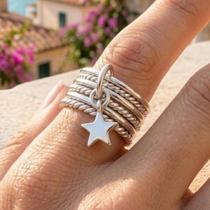 May include: A silver ring set with multiple bands, some twisted, and a dangling star charm. The star is white and has a five-point design. The ring is worn on a finger.