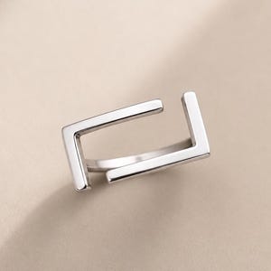 Könnte beinhalten: Ein minimalistischer Silberring mit offenem, geometrischem Design. Der Ring hat eine quadratische Form mit zwei offenen Enden, was einen modernen und stilvollen Look erzeugt. Das polierte Metall reflektiert das Licht und unterstreicht die klaren Linien und die moderne Ästhetik.