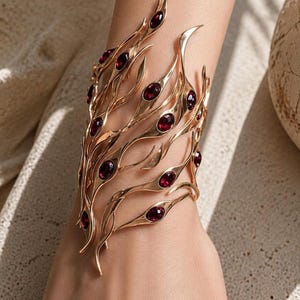May include: A gold-coloured cuff bracelet with a fluid, organic design. The bracelet features multiple curved, leaf-like elements set with oval, deep red gemstones. The jewellery piece is worn on a wrist, showcasing its intricate details and elegant style.