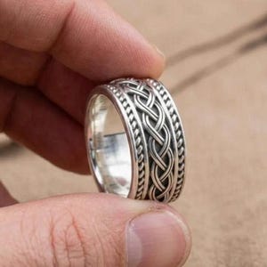 May include: A silver ring with a Celtic knot design. The ring has a braided pattern in the centre, framed by small, textured details. The ring is held by a person's fingers.