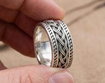 May include: A silver-colored ring with a Celtic knot design. The ring features a braided pattern in the center, bordered by a series of small, textured details. The ring is held up by a person's fingers.