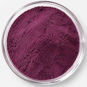 May include: A clear glass bowl filled with a fine, deep purple powder. The powder has a matte, textured surface with subtle variations in density and small clumps, contained within the circular rim of the transparent glass vessel.