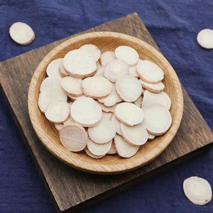 May include: A wooden bowl containing many thinly sliced, pale-coloured pieces, possibly a root vegetable. The bowl rests on a dark wooden board against a blue fabric backdrop. Several slices are scattered around the bowl.