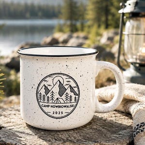 May include: A white ceramic mug with a black speckled finish and a black rim. The front features a circular black graphic of mountains, pine trees, and a tent with the text "CAMP HOWBIOWALSKI" and "2025" printed in a clean, sans-serif font.
