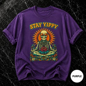 Puede incluir: Camiseta morada con el texto "STAY YIPPY" sobre un esqueleto con gafas de sol, sentado en posición de loto. El esqueleto está rodeado por un diseño de sol y elementos florales. La camiseta tiene las mangas enrolladas.