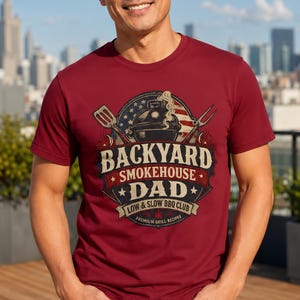 Pode incluir: Uma camiseta bordô com um gráfico temático de churrasco. O design inclui uma grelha, estrelas e o texto "Backyard Smokehouse Dad, Low & Slow BBQ Club, Premium Grill Recipes". A camiseta é de manga curta.