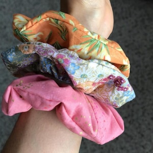 May include: A stack of three fabric scrunchies in various floral patterns. The top scrunchie is orange with green leaves, the middle one has blue and purple flowers, and the bottom one is pink with gold flecks.