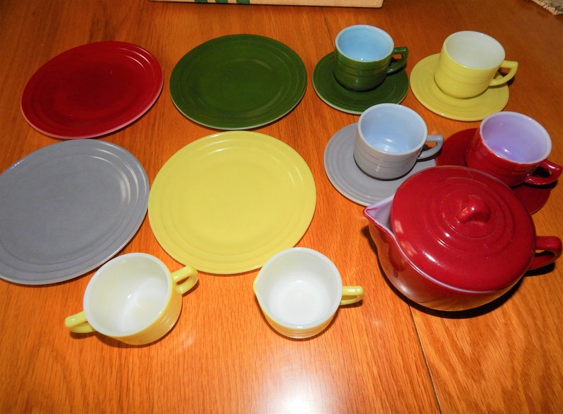May include: A vintage children's tea set with a red teapot, yellow, green, and gray plates, and cups in various colors, including yellow, blue, and purple.