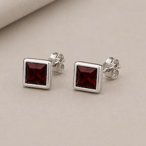 May include: A pair of silver stud earrings featuring square-cut, deep red garnet gemstones set in a smooth, polished silver frame. Each earring includes a standard post back mechanism for secure attachment, showcasing a clean, geometric design.
