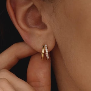 May include: A gold hoop earring featuring a double-band design. One band is a smooth, polished gold surface, while the adjacent band is set with a row of small, clear, sparkling stones that catch the light, creating a layered appearance.