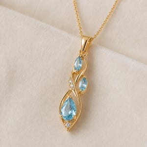 May include: A gold pendant necklace featuring a vertical arrangement of three light blue gemstones. The bottom stone is pear-shaped, while the two upper stones are marquise-cut. Small clear accent stones are set within the gold metal frame.