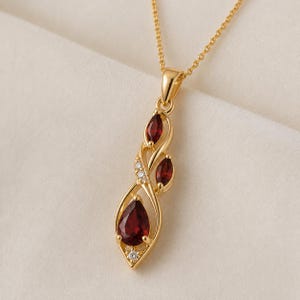 May include: A gold pendant necklace featuring a vertical arrangement of three deep red garnet gemstones in pear and marquise cuts, accented by two small clear round stones, all set within a polished gold frame suspended from a delicate gold chain.