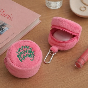 Può includere: Due custodie rosa in peluche, una rotonda con testo verde ricamato "Happy Birthday Harry". L'altra è aperta, con cerniera e moschettone argentato. È visibile anche un rossetto rosa.
