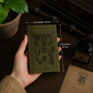 May include: A green pocket book titled "The Practical Pocket Book of Herbology," measuring 3.9 inches wide and 5.9 inches tall. The cover features botanical illustrations. The book is held near a potted plant, inkwell, and a letter with the Hogwarts crest.