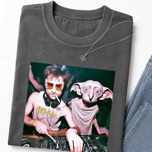 May include: Dark gray t-shirt featuring a graphic of a DJ with orange sunglasses and a creature. The shirt has a crew neck and short sleeves. A silver necklace with a sun pendant is on the shirt. Light blue jeans are visible.