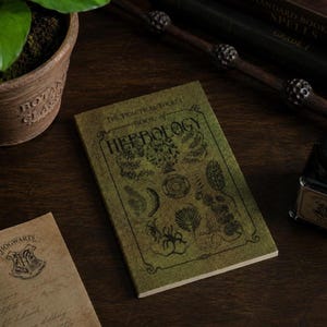 May include: A small, green book titled "The Practical Pocket Book of Herbology" with botanical illustrations. The book is on a dark wooden surface, with a wand and other items in the background. The book is a prop from the Harry Potter series.