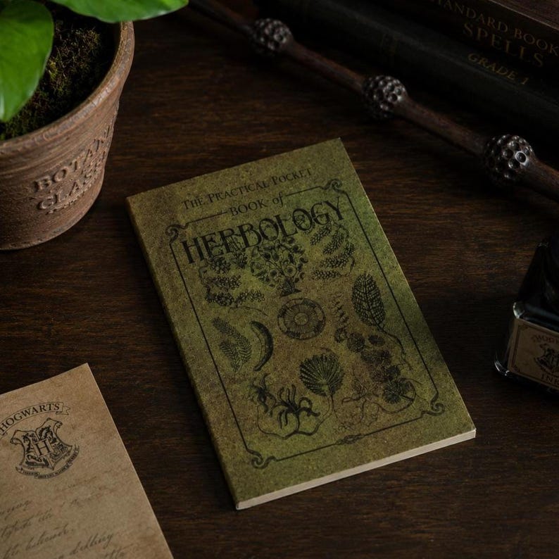 May include: A small, green book titled "The Practical Pocket Book of Herbology" with botanical illustrations. The book is on a dark wooden surface, with a wand and other items in the background. The book is a prop from the Harry Potter series.