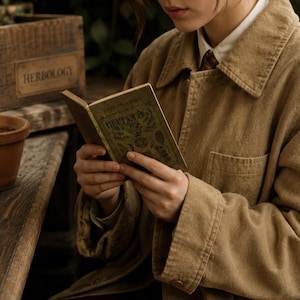 May include: A person reading a small, antique book titled "Herbology." The book has a green cover with gold lettering. The person is wearing a tan jacket and a white collared shirt. A wooden box with "Herbology" printed on it is in the background.