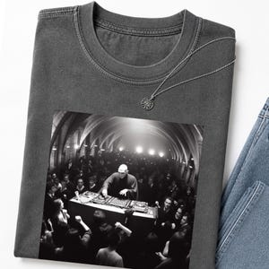 Può includere: T-shirt grigio scuro con un'immagine in bianco e nero di un DJ che si esibisce davanti a una folla. Una collana d'argento con un ciondolo circolare è sulla maglietta. La maglietta è abbinata a jeans blu.