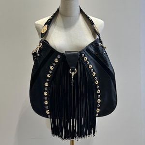 May include: A black hobo bag featuring long fringe detailing on the front center, accented by rows of gold-tone metal studs along the sides. The bag includes a wide shoulder strap with gold-tone hardware and a central clasp closure on the front.