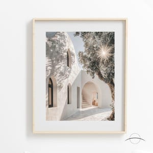 May include: Framed print featuring a sunlit architectural scene. A white building with arched doorways and windows is visible, with shadows cast by a tree. The print is in a light wood frame, creating a minimalist aesthetic.
