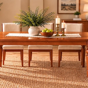 May include: An oval wooden dining table with four turned legs, featuring a white table runner, a white speckled vase with green foliage, a wooden bowl filled with green apples, and two brass candlesticks with white pillar candles.