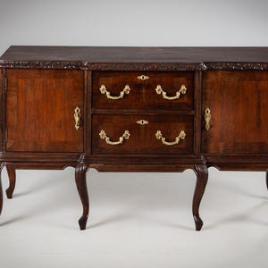 Puede incluir: Aparador de madera marrón oscuro con dos puertas y dos cajones. Las puertas y los cajones tienen tiradores y cerraduras doradas. El aparador tiene tallas ornamentadas en la parte superior y patas curvas.