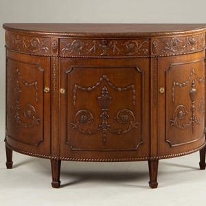 Puede incluir: Un aparador de madera en forma de media luna con detalles tallados de flores y volutas. El mueble tiene tres puertas con tallas ornamentadas y herrajes dorados. El diseño curvo está sostenido por cuatro patas cortas. La madera tiene un acabado marrón cálido.