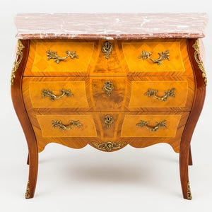 May include: A wooden chest of drawers with a marble top. The chest has a curved front with four drawers, each with ornate brass handles. The wood has a warm, golden hue with intricate patterns and detailing.
