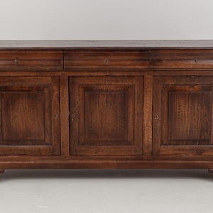 Puede incluir: Aparador de madera marrón oscuro con tres cajones y tres puertas de armario. Los cajones tienen pequeños agujeros para llaves y las puertas del armario tienen paneles empotrados. El aparador es un mueble clásico.