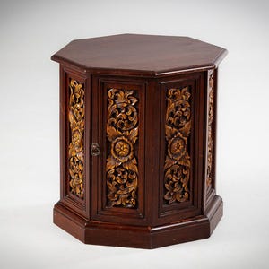 Puede incluir: Un mueble de madera hexagonal de color marrón oscuro con intrincadas tallas florales. El mueble tiene una pequeña puerta con un tirador de metal y es adecuado para el almacenamiento. Los detalles tallados se destacan con un tono dorado más claro.