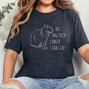 May include: Heather dark grey t-shirt with a white outline drawing of a capybara and the text "IDK HOW MUCH LONGER I CAN SLAY". The shirt is worn by a person sitting on a white couch.