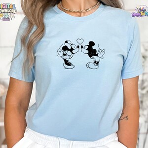 May include: A light blue short-sleeve t-shirt featuring a black graphic print of two cartoon mice kissing with a small heart positioned between them. The shirt has a crew neckline and a simple, clean design on the front center of the garment.