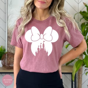 May include: Heather mauve t-shirt featuring a white Minnie Mouse bow and silhouette of a castle. The shirt has short sleeves and a round neckline. The text "Heather Mauve" is in a pink circle in the bottom left corner.