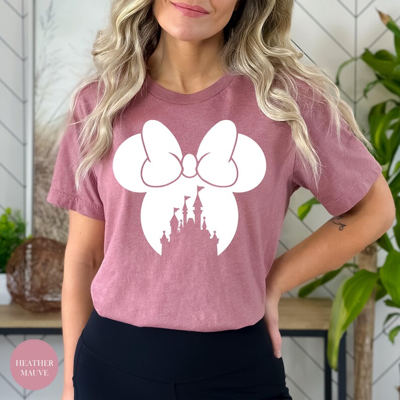 May include: Heather mauve t-shirt featuring a white Minnie Mouse bow and silhouette of a castle. The shirt has short sleeves and a round neckline. The text "Heather Mauve" is in a pink circle in the bottom left corner.