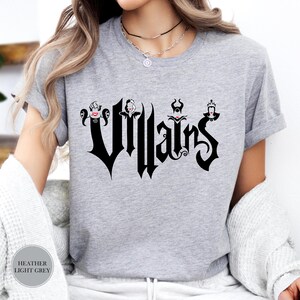 May include: Heather light grey t-shirt featuring the word "Villains" in a gothic font, with stylized character illustrations above the letters. The shirt is a light grey color.