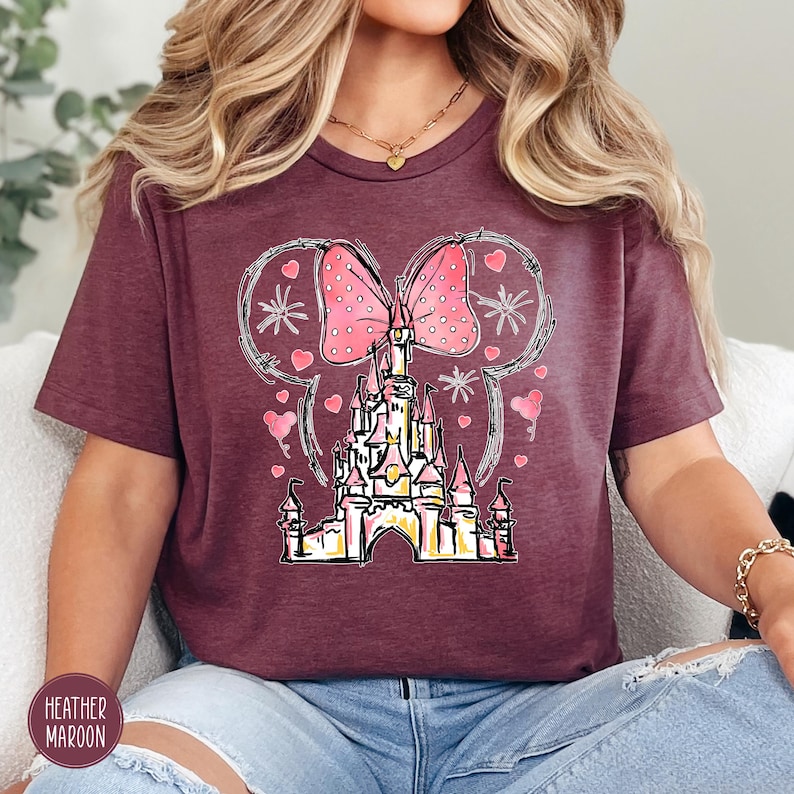 Puede incluir: Camiseta granate con un dise&ntilde;o de castillo caprichoso, un lazo rosa y detalles de corazones. El dise&ntilde;o incluye un castillo, corazones y orejas de Mickey Mouse. El color de la camiseta es un rojo-p&uacute;rpura intenso.