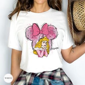 May include: White t-shirt with a pink Minnie Mouse-shaped design, a polka dot bow, and a princess illustration. The word "Aurora" is written in cursive. The shirt has a classic crew neck.