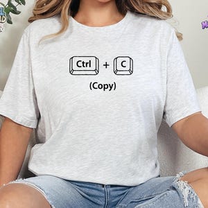 May include: A light gray short-sleeved t-shirt featuring a black graphic of two computer keyboard keys labeled Ctrl and C with a plus sign between them and the word (Copy) printed in black text underneath the keys on the front of the shirt.