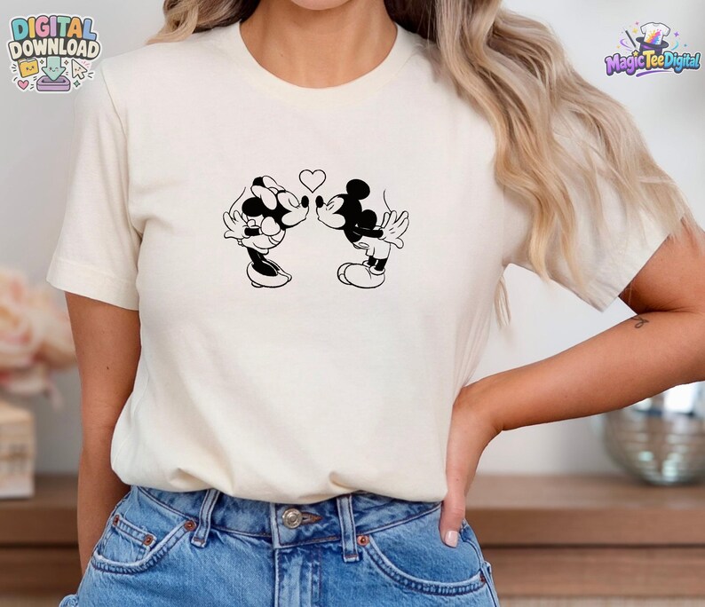 May include: A cream-colored short-sleeve t-shirt featuring a black line art graphic of two cartoon mice kissing with a small heart positioned between them. The design is centered on the chest area of the shirt, which is paired with blue denim jeans.
